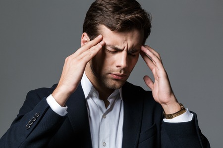 Close up portrait of a exhausted young businessman dressed in suit isolated over gray background, suffering from a headacheの写真素材