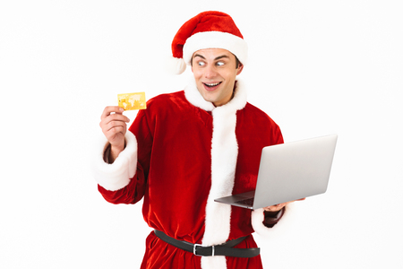 Portrait of an excited young man dressed in santa claus costume standing isolated over white background, holding laptop computer, showing plastic credit cardの写真素材