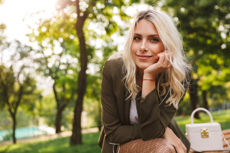 Image of adorable woman 20s smiling at camera while sitting on bench in park with treesの写真素材