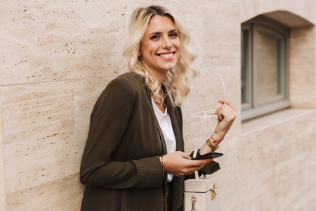 Happy young woman leaning on a wall at the city, holding mobile phoneの写真素材
