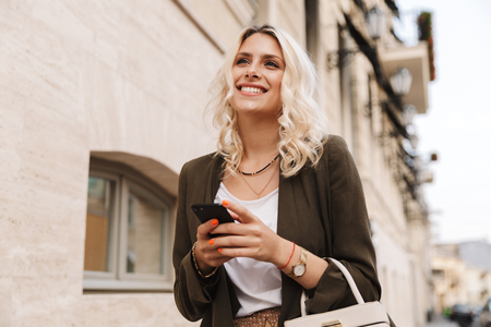 Image of caucasian woman wearing jacket using mobile phone for chat while walking through city streetの写真素材