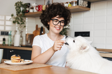 Image of a pretty curly woman sitting at the kitchen eat cake and feed her cutie white dog.の写真素材