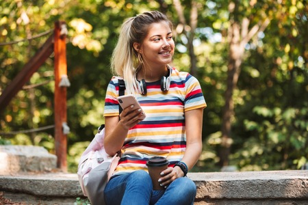 Image of a happy cute young girl using mobile phone holding coffee.