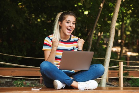 Portrait of an excited young teenage girl sitting on a bench at the park, using laptop computer on her lap, celebratingの写真素材