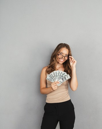 Portrait of a confident young business woman standing isolated over gray background, showing money banknotesの写真素材
