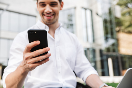 Close up of happy young business man using mobile phone while sitting outdoors with laptop computerの写真素材