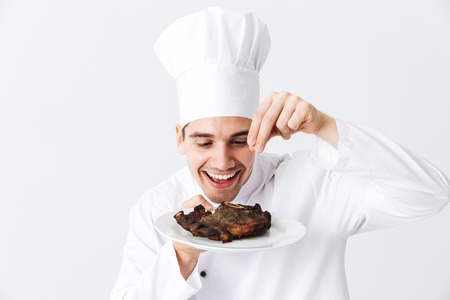 Cheerful chef cook wearing uniform peppers cooked beef steak on a plate isolated over white...