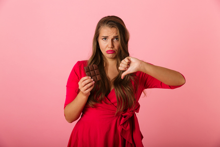 Photo of a pretty sad young woman isolated over pink wall background holding chocolate showing thumbs down.の写真素材
