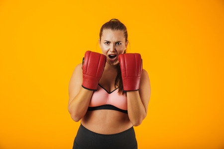 Portrait of beautiful big woman in sportive bra wearing boxing gloves practicing isolated over yellow backgroundの写真素材