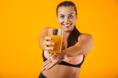 Image of caucasian chubby woman in tracksuit smiling and holding glass with orange juice isolated over yellow backgroundの写真素材