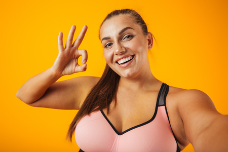 Portrait of cheerful chubby woman in sportive bra using mobile phone for selfie photo isolated over yellow backgroundの写真素材