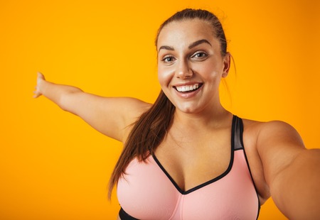 Portrait of a cheerful overweight fitness woman wearing sports clothing standing isolated over yellow background, taking a selfie with mobile phoneの写真素材