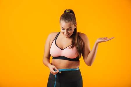Portrait of a cheerful overweight fitness woman wearing sports clothing standing isolated over yellow background, holding measure tape over her waistの写真素材
