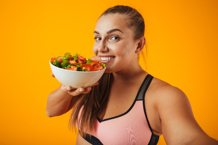 Portrait of a cheerful overweight fitness woman wearing sports clothing standing isolated over yellow background, taking a selfie with mobile phone, showing bowl with saladの写真素材