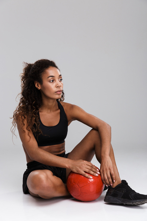 Portrait of an afro american confident young fit sportswoman doing exercises with heavy ball isolated over gray background, sitting, looking awayの写真素材