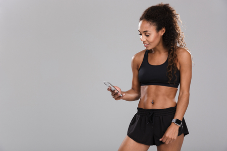 Beautiful african sportswoman isolated over gray background, using mobile phoneの写真素材