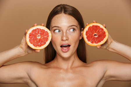Beauty portrait of a beautiful healthy young topless woman with long red hair showing sliced grapefruit isolated over beige backgroundの写真素材