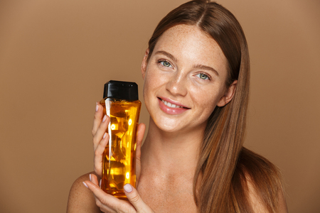 Beauty portrait of a smiling young topless woman with long red hair showing bottle of shampoo isolated over beige backgroundの写真素材