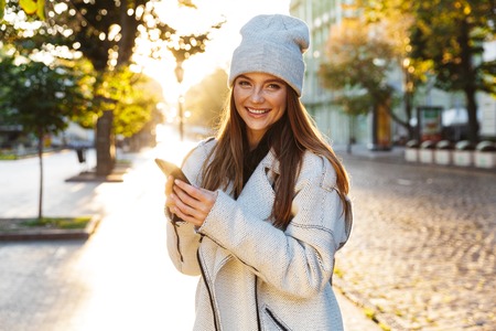 Beautiful young woman dressed in autumn coat and hat walking outdoors, using mobile phone