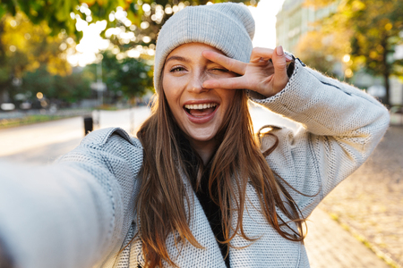 Beautiful young woman dressed in autumn coat and hat walking outdoors, taking a selfieの写真素材
