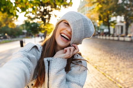 Beautiful young woman dressed in autumn coat and hat walking outdoors, taking a selfieの写真素材