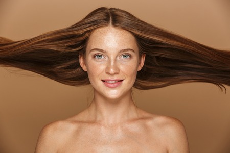 Beauty portrait of a beautiful healthy young topless woman with long red hair looking at camera isolated over beige backgroundの写真素材