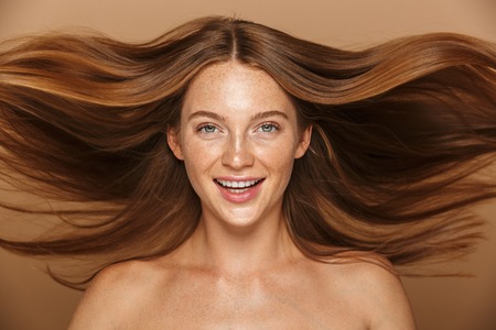 Beauty portrait of a beautiful healthy young topless woman with long red hair looking at camera isolated over beige backgroundの写真素材