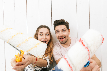 Image of european couple man and woman holding paint rollers and standing over white wall while painting and making renovationの写真素材