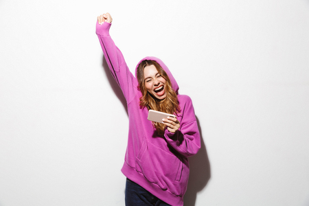 Portrait of an excited girl wearing hoodie standing isolated over white background, celebrating success, holding mobile phoneの写真素材