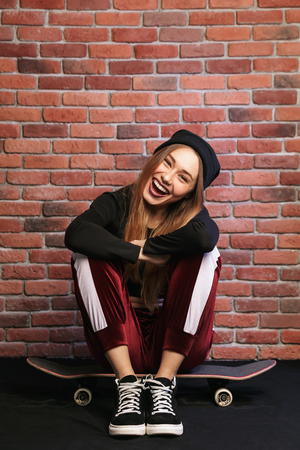 Image of pretty sporty girl 20s laughing while sitting on skateboard against brick wallの写真素材
