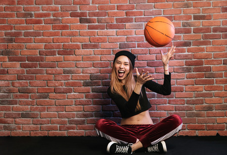 Photo of artistic girl 20s sitting on floor against brick wall and playing with basketballの写真素材