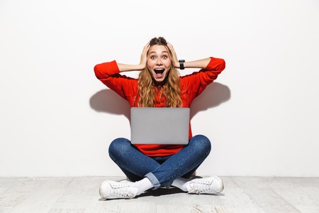 Portrait of an excited young woman wearing hoodie sitting isolated over white background, using laptop computer, celebrating successの写真素材