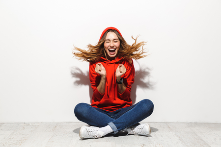 Portrait of an excited girl wearing hoodie sitting on a floor isolated over white backgroundの写真素材