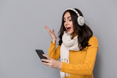 Portrait of excited woman wearing ear muffs and scarf holding mobile phone isolated over gray backgroundの写真素材