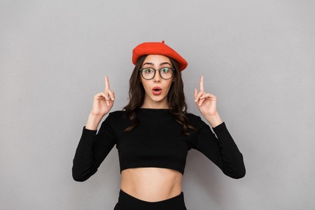 Portrait of an excited young woman dressed in red hat standing over gray background, pointing upの写真素材