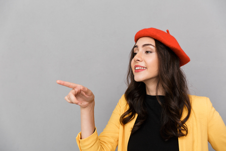 Close up of a smiling young woman in red hat standing over gray background, pointing finger away at copy spaceの写真素材