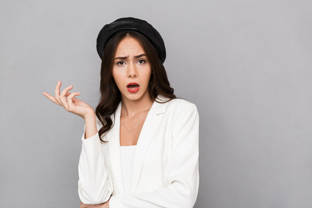 Portrait of a confused young woman wearing beret standing isolated over gray background, looking at cameraの写真素材