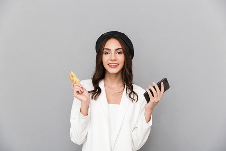 Portrait of a happy young woman dressed in jacket over gray background, holding mobile phone,...