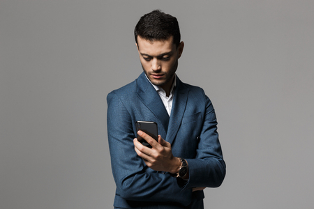Image of serious concentrated business man holding mobile phone posing isolated over grey wall background.の写真素材