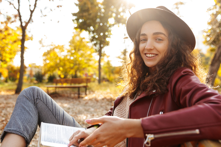 Image of an amazing cheerful beautiful woman sitting on a bench in park reading book.の写真素材