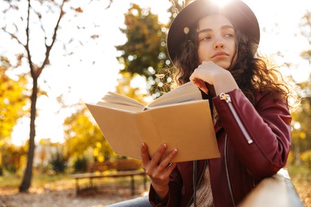 Image of a beautiful cute woman sitting on a bench in park reading book.の写真素材