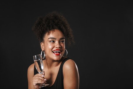 Portrait of a beautiful afro american woman wearing makeup standing isolated over black background, holding glass of champagneの写真素材