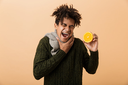 Portrait of sick african american guy wearing sweater and scarf touching throat and holding half an orange isolated over beige backgroundの写真素材