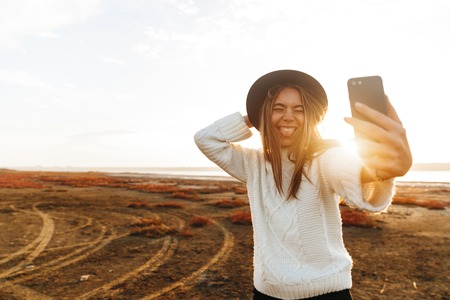 Beautiful smiling young woman walking outdoors at the beach during sunset, taking a selfieの写真素材