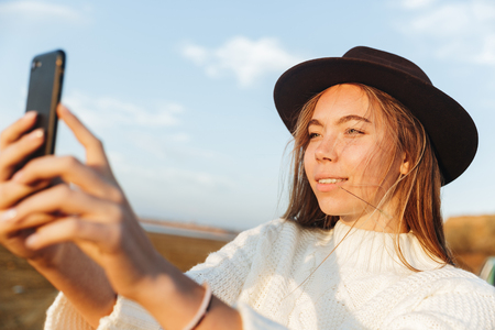 Beautiful smiling young woman walking outdoors at the beach during sunset, taking a selfieの写真素材