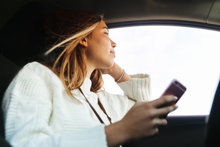 Beautiful young woman holding mobile phone while sitting in a car, looking at windowの写真素材