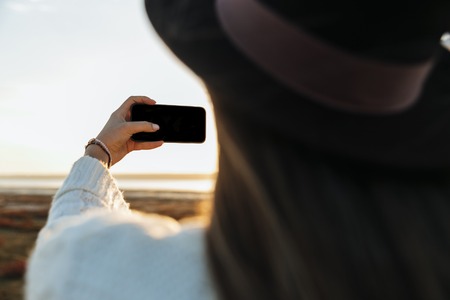 Back view of a young woman walking outdoors at the beach during sunset, taking a selfieの写真素材