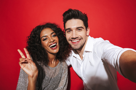 Portrait of a cheerful young smartly dressed couple standing isolated over red background, taking a selfie with outstretched handの写真素材