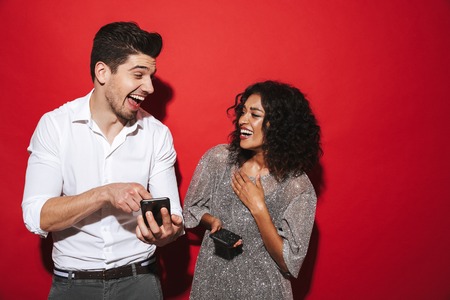 Portrait of a cheerful young smartly dressed couple standing isolated over red background, holding...