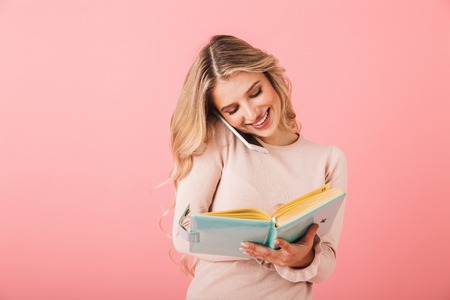 Portrait of a cheerful young woman wearing sweater standing isolated over pink background, writing notes in a diary while talking on mobile phoneの写真素材